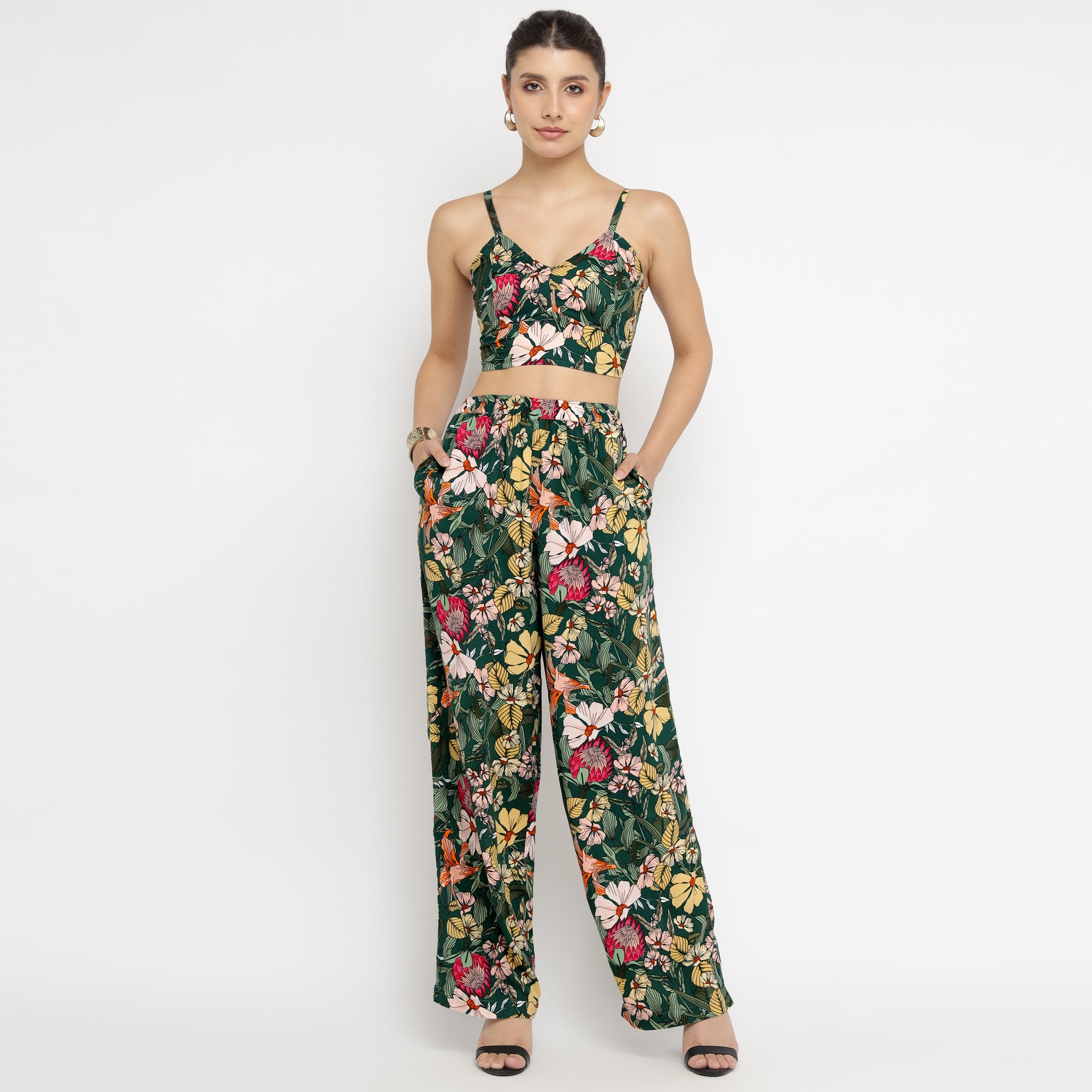 Front view of a model wearing the Tropical Bloom Co-ord Set, featuring a strappy crop top and high-waisted wide-leg pants with a vibrant floral print in green, yellow, pink, and red hues. The model is standing against a white backdrop with her hands in the pants’ pockets, displaying the comfortable fit and bold tropical pattern of the outfit.